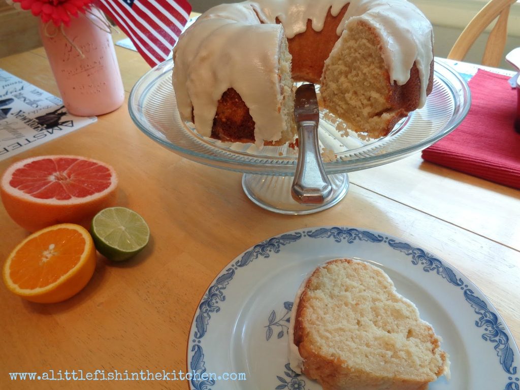 A freshly baked vanilla buttermilk pound cake is is served on a clear glass cake stand. The cake is cut revealing a golden crumb on the interior of the cake. A thick, rich citrus glaze is drizzled over the top of the cake making it look sweet and tempting. A butter knife rests the cake plate and a blue ceramic dessert plate is just below the cake plate with a single slice of iced cake on it, ready to enjoy. Just beside the cake plate, one half of each type of citrus fruit used in the simple glaze are on the table. The colorful insides of an orange, a pink grapefruit and one green lime are on the table for a colorful decorative effect.