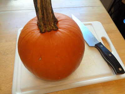 A small, whole pumpkin is on a white plastic cutting board with a sharp chef's knife beside it. 