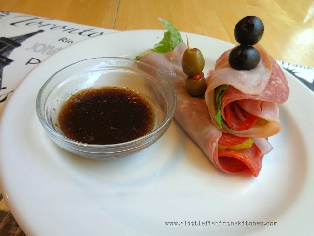 Two low-carb Italian sub rolls up are ready to be enjoyed. Layers of flavorful, thinly sliced ham, salami, red onion, pepperoncini peppers and Romaine lettuce are all descernable from the sides of the roll ups. The rolls ups are pinned together with a toothpick garnished with black and green  olives. The white serving plate also has a small clear glass ramekin with homemade balsamic dressing ready for dipping. 