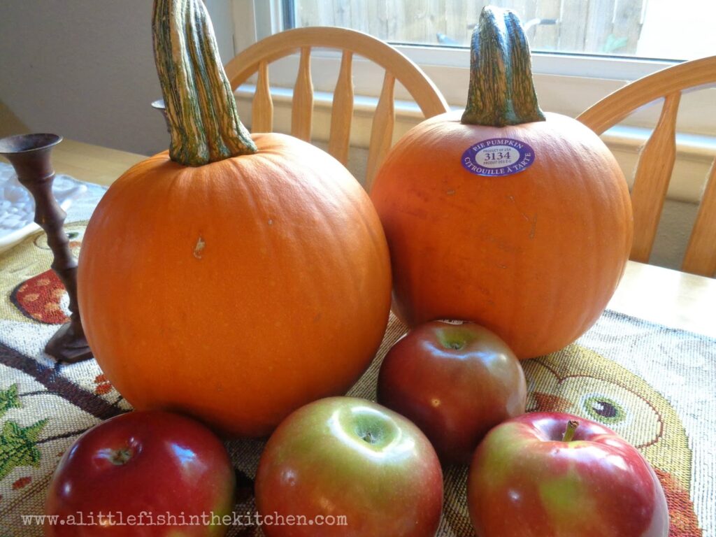 Two fresh, petite pie pumpkins are on display with fresh apples. The fresh pumpkins and apples are next to a sunny window on a wooden table top with a fall theme table runner. The runner has embroidered owls and colorful leaves.