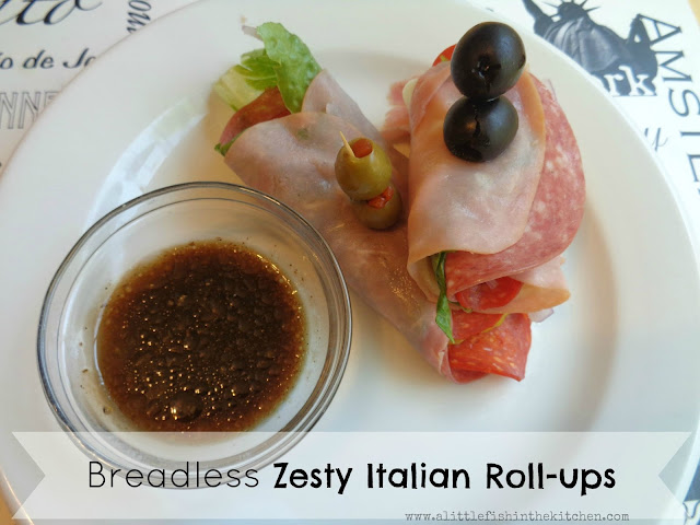 Two low-carb Italian sub rolls up are ready to be enjoyed. Layers of flavorful, thinly sliced ham, salami, red onion, pepperoncini peppers and Romaine lettuce are all descernable from the sides of the roll ups. The rolls ups are pinned together with a toothpick garnished with black and green  olives. The white serving plate also has a small clear glass ramekin with homemade balsamic dressing ready for dipping.