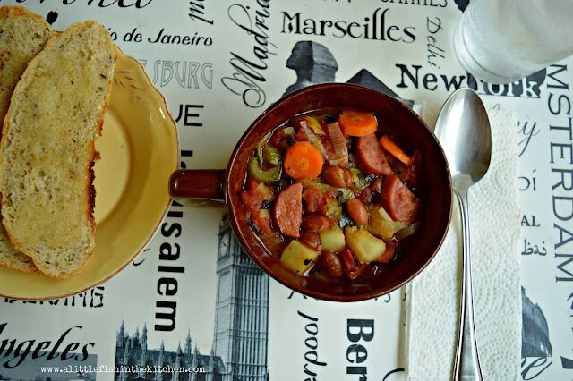 Hearty smoked sausage stew is served in a vintage brown ceramic soup bowl with a handle. Tender sliced carrots, diced potato, celery, leeks, half-moon shaped smoked sausge pieces, and whole pinto beans are visible in this hearty stew. The bowl is on  a white place mat decorated with black words and a spoon rests just beside the bowl. 