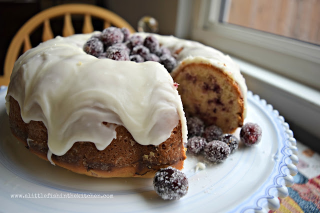 A festively beautiful cranberry orange pound cake is ready to serve on a blue and white marbled cake stand. The cake is decorated with sugared whole cranberrys in the center that have scattered onto the cake plate after the cake was sliceed. The crumb is lighly golden and bits of chopped cranberry, orange zest and visible in the crumb. The white glaze look rich and sweet drizzled over the sides of the cake. 