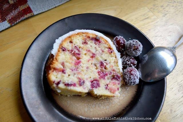 A single slice of rich buttery cranberry pound cake is served on a small, brown ceramic plate. The golden crumb of the cake slices is dotted with fresh bits of cranberry and orange zest. The white icing looks thick and inviting. The cake is garnished with two sugared cranberries on the side. 