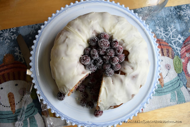 The top of a freshly-iced cranberry orange pound cake with the cavity filled with festively sugared whole cranberries. One slice is removed and the whole cranberries have tumbled into the cut. The cake is on a blue and while marbled caked stand which is on a festive, sparkly blue and white table runner that's decorated with snowmen wearing scarves. 