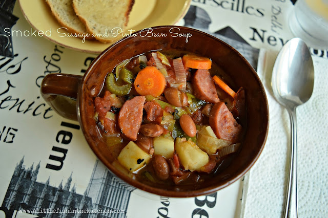 Hearty smoked sausage stew is served in a vintage brown ceramic soup bowl with a handle. Tender sliced carrots, diced potato, celery, leeks, half-moon shaped smoked sausge pieces, and whole pinto beans are visible in this hearty stew. The bowl is on  a white place mat decorated with black words and a spoon rests just beside the bowl. 