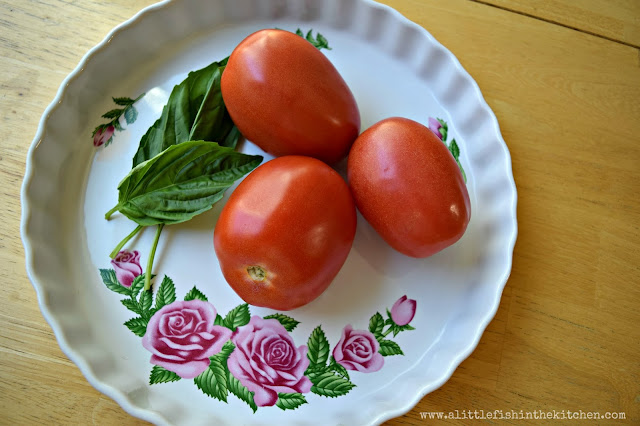 Three red, rip Roma tomatoes and whole fresh basil leaves are featured laying inside of a vintage white ceramic tart dish. The tart dish has a pattern of pink roses and green leaves in the ceramic coating.
