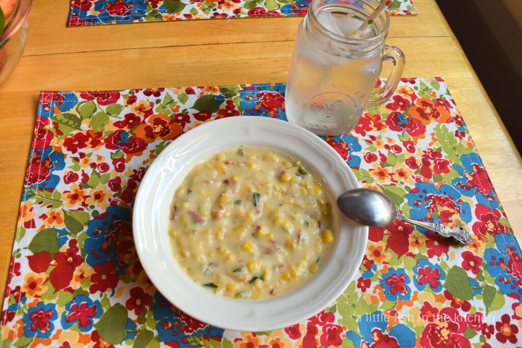 A bowl of colorful corn chowder sits on a colorful placemat decorated with a primary colored floral print. The print has bright blues, red, orange, yellow and spring green. The creamy corn chowder is has a yellow hue and bit of corn, green pepper, bacon, onion and herbs and spices are visible in the hearty soup. 