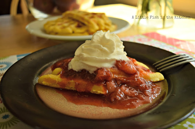 A plate of warm waffles is ready to serve with prepared strawberry jam. In the center of the tabletop, a single plate with a waffle topped with rich, red strawberry jam and whipped cream is ready to be enjoyed. Bits of whole strawberries are visible in the richly-colored, homemade jam.