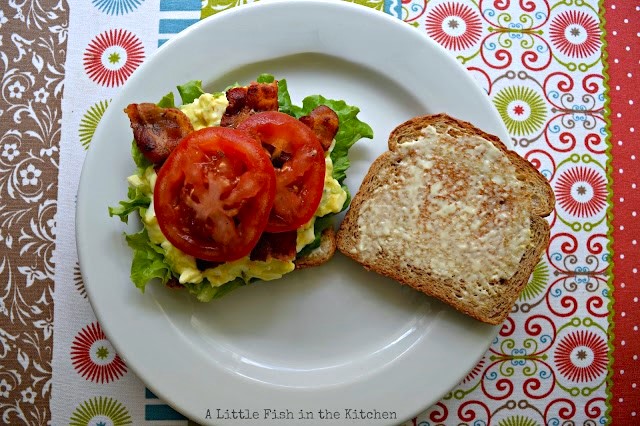 An inviting open face egg salad BLT is on toasted bread. It's on a white plate and ready enjoy. The layers of crisp green lettuce, yellow egg salad, crispy cooked bacon and ripe tomatoes are eye-catching.
