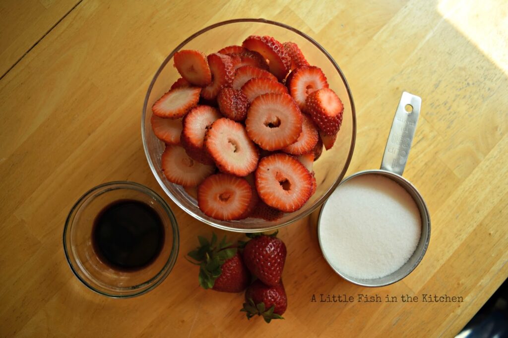 One pound of fresh, ripe strawberries are in a glass bowl, a measuring cup with sugar, and a ramekin with a measure of balsamic vinegar are ready to use. 