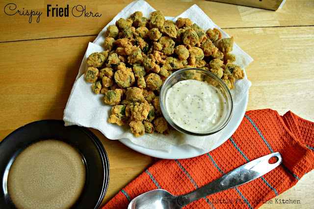 A platter filled with fresh crispy fried okra is read to serve. A clear glass ramekin with Southern cream gravy is nestled in the pile of breaded okra.