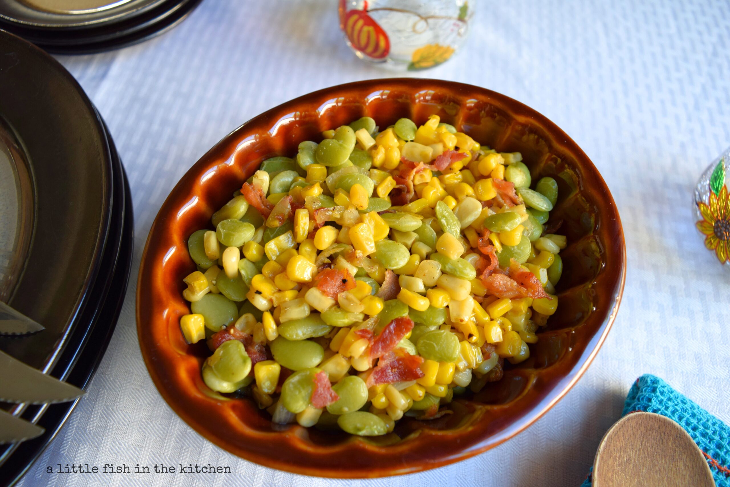 A colorful medley of corn, Lima beans and bacon is ready to serve in a brown ceramic bowl. The bowl is on a table draped with a white tablescloth and warmly colored fall decor, pumpkins and sunflower-theme items are slightly blurred in the background. 