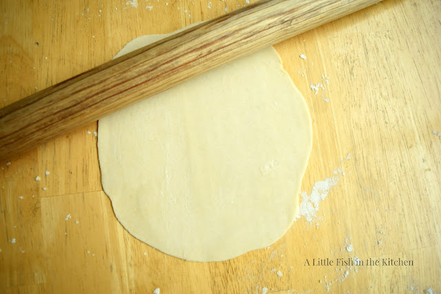 A single tortilla is rolled out into a circular form on a wooden tabletop and is ready to cook. The wooden French rolling pin is next to the prepared tortilla. 