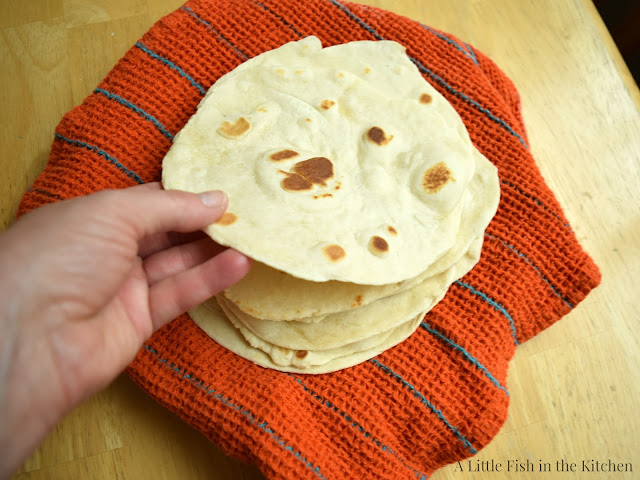 A hand is lifting a single freshly prepared tortilla from a stack of homemade flour tortillas. 
