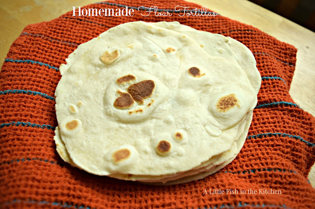 Fresh, homemade flour tortillas are wrapped in a cotton tea towel to stay warm. The tortillas look soft and are perfectly speckled with brown griddle marks. The tea towel or burnt orange with teal blue stripes. 