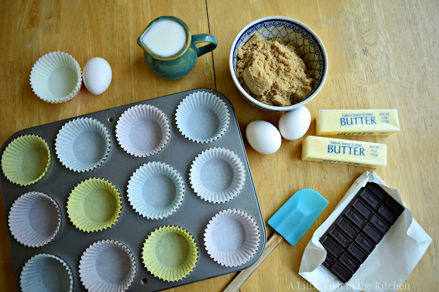 A muffin tin lined with paper liners is surround with all the ingredients to make chocolate cupcake from scratch. A bar of baking chocolate, butter, eggs, brown sugar, and milk.