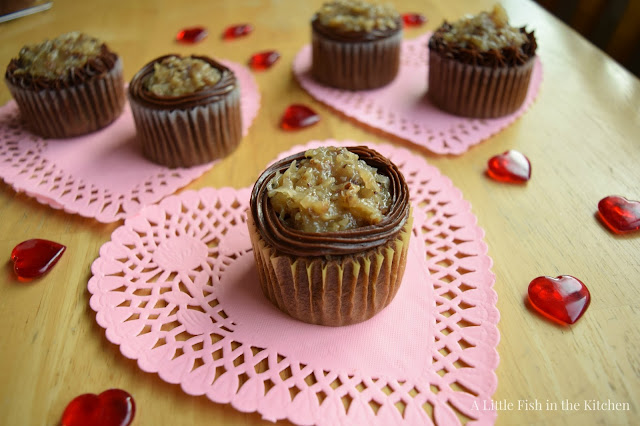 German chocolate cupcakes are topped with homemade coconut pecan icing, and accented with piped borders of chocolate icing. The cupcakes are on pink paper doilies with decorative glass hearts spread around the table.
