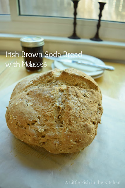 A fresh baked loaf of rustic, freeform Irish brown soda bread is cooling on a wooden table top by a large window. Two white bread plates, a butter knife and a mason jar filled with homemade strawberry jam are beside the bread for serving. 