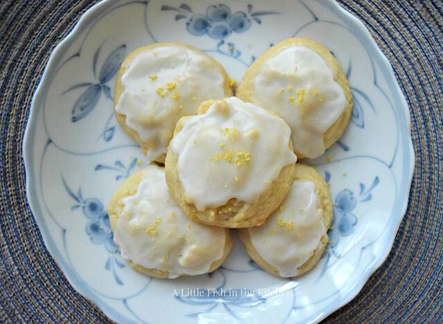 A small stack of tempting iced lemon cookies is on a vintage-style white plate with a blue floral pattern. The cookies appear soft with a mild yellow hue. Bits of fresh, bright yellow lemon zest garnish the tops of the cookies. 