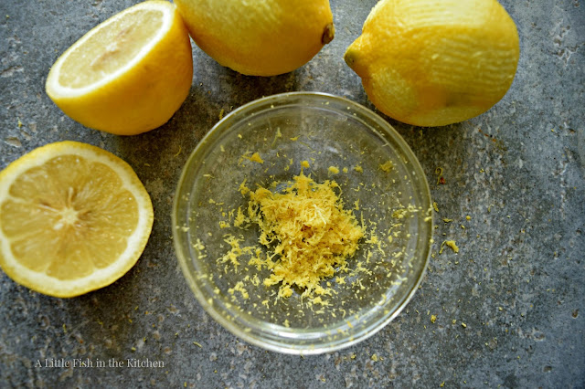 A clear glass ramekin holds a few tablespoons of yellow, fresh lemon zest. Whole freshly zested lemons are next to the small bowl. The zesting marks are visible in the fruit skins. 
