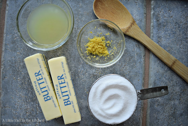 Clear glass ramekins with lemon juice, lemon zest, a one cup measuring cup filled with sugar, two sticks of butter and a wooden spoon are set out to bake lemon sugar cookies. 