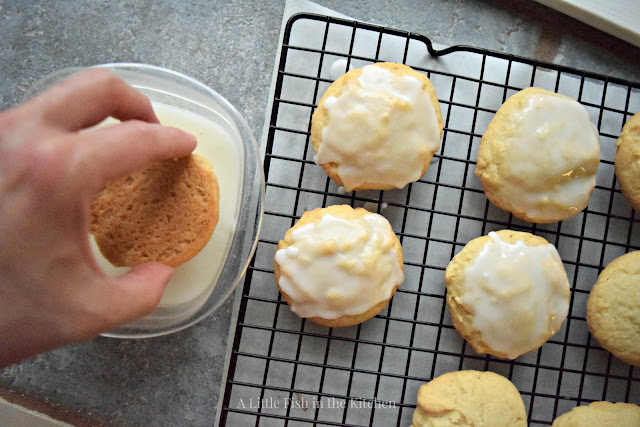 Soft lemon cookies are iced by dipping the tops of the cookies in a light, sweet lemon glaze. The fresh baked cookies are iced and draining on a rack placed over a baking sheet. 