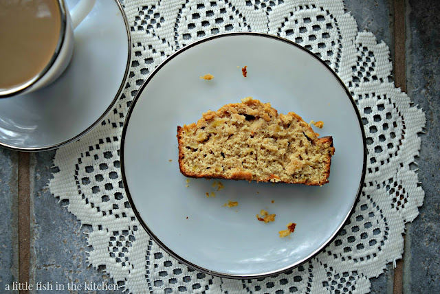 A single slice of this simple apple quick bread is served on a vintage milk glass plate with a silver rim. The plate is resting on a vintage white lace doillie. A cup of coffee with cream is served in a vintage white milk glass cup and saucer set. The slice of fresh apple bread look spiced, tender and tempting with bits of fresh apple speckled throughout the crumb.  