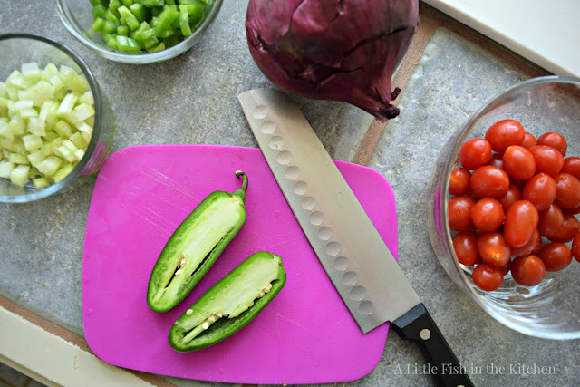Fresh ingredients gathered and being prepared for this flavorful corn salad. A set of small clear glass bowls filled with chopped fresh green peppers, chopped celery, whole cherry tomatoes surround a small purple plastic cutting board. A fresh jalapeno pepper is sliced in half on top of the cutting board and whole fresh purple onion sits beside the cutting board. 
