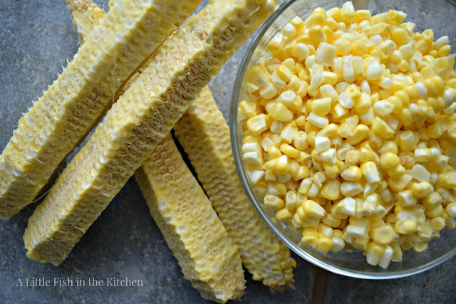 Four raw corn husks rest near a clear glass bowl filled with their fresh corn kernels. The kernels are freshly scraped from the cob and delightfully light yellow.