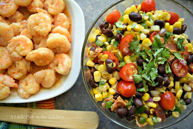 A richly-colored corn and black bean salad sits in a clear glass serving bowl. A green and blue plaid tea towel sits near to the bowl with a wooden spoon resting on top. A platter of grilled shrimp sits just beside the bowl of corn salad, the meal is ready to serve. The corn and black bean salad has brightly colored ingredients that include yellow corn, red ripe cherry tomatoes, green fresh peppers, bacon bits and hearty black beans.