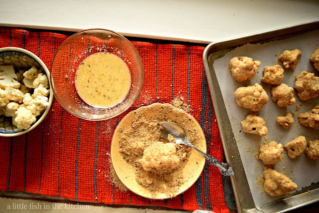 An assembly line for making homemade oven fried cauliflower. Separate bowls filled with fresh cauliflower florets, egg wash, and homemade bread crumb coating are set out beside a rimmed baking dish. The baking dish is prepped with a sheet of parchment paper. Crumb -coated cauliflower florets are ready for the oven. 