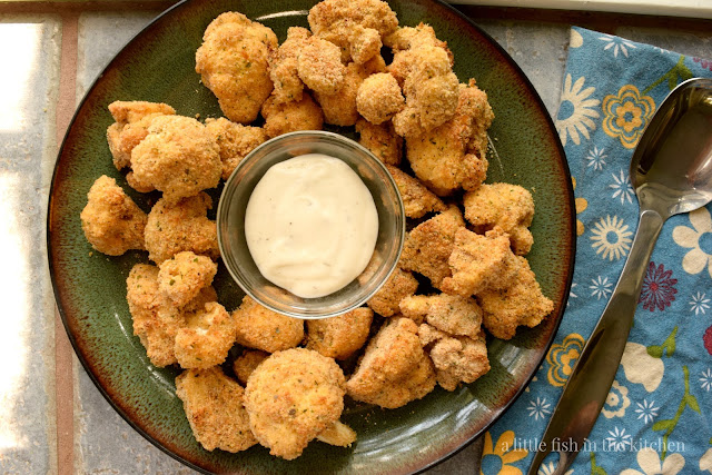 Crispy breaded fried cauliflower florets are arranged on a dark green ceramanic platter surrounding a small ramekin of ranch dressing. A bright blue cotton tea towel with a bright floral patterns rests just beside the plate. A silver metal serving spoon rests on top of the towel. 