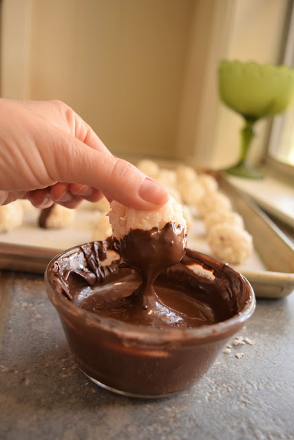 A woman's hand is dipping a no-bake coconut candy balls into melted chocolate as the final step to make vintage coconut joy candies with almonds. 