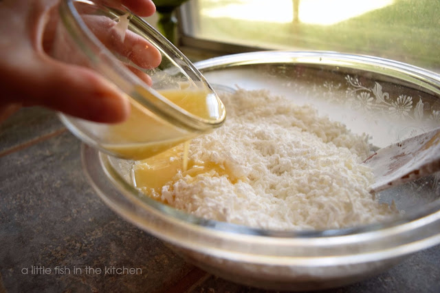  Melted butter is being poured in the a mixture of sweetened coconut flakes and powdered sugar. The clear glass mixing bowl is near a sunny kitchen window. 