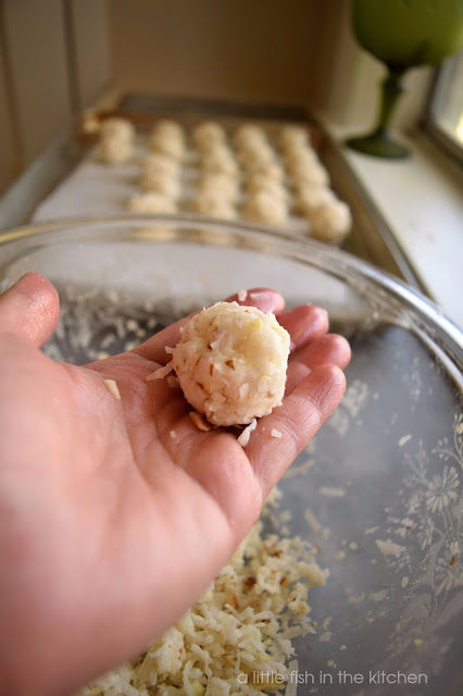 The moistened coconut mixture is rolled into candy balls by hand. The coconut candies are then places on a baking sheet lined with parchment paper to rest until being dipped in melted chocolate. A close up shot of a woman's hand holding a freshly formed coconut candy ball is front and center in the photo. 
