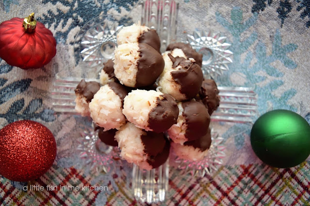 Tempting chocolate-covered coconut candies speckled with toasted almond bits are arranged on a clear glass vintage plate. The homemade Coconut Joys have a soft, chewy coconut center and a glossy chocolate coating. The plate is on a winter-themed table runner with blue and white snow flakes and a plaid edge. 3 vintage-style, red and green Christmas ornaments surround the plate for a decorative effect. 