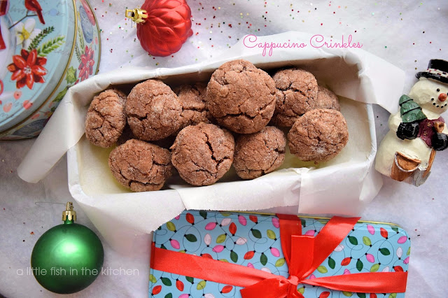 One dozen chocolate cappuccino crinkle cookies are in a Christmas themed metal treat box. The box is light teal with a jumbled pattern of Christmas string lights that are pink, white, dark and light green. The metal gift box also has a red bow attached. The cookies have a rich, brown color and delightfully crackled tops. 