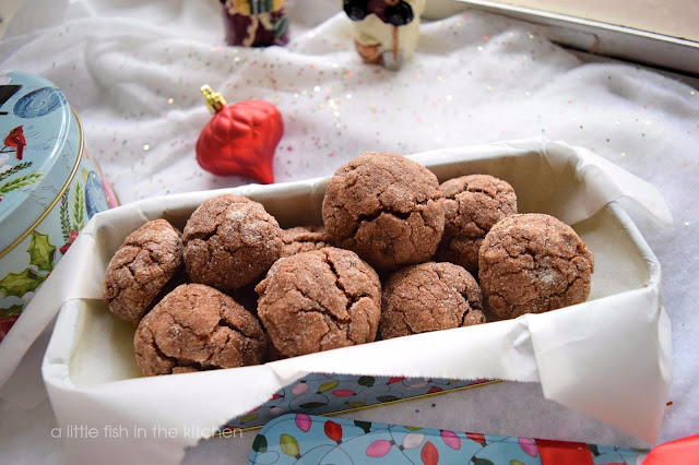 One dozen chocolate cappuccino crinkle cookies are in a Christmas themed metal treat box. The box is light teal with a jumbled pattern of Christmas string lights that are pink, white, dark and light green. The metal gift box also has a red bow attached. The cookies have a rich, brown color and delightfully crackled tops. 