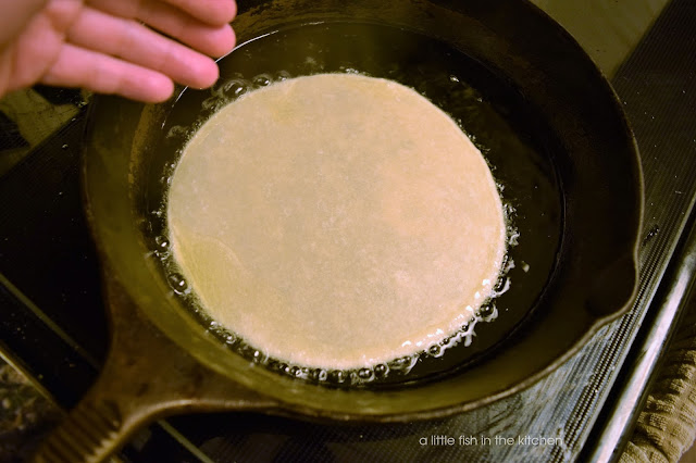 An uncooked tortilla is dropped into hot oil in a cast iron skillet.