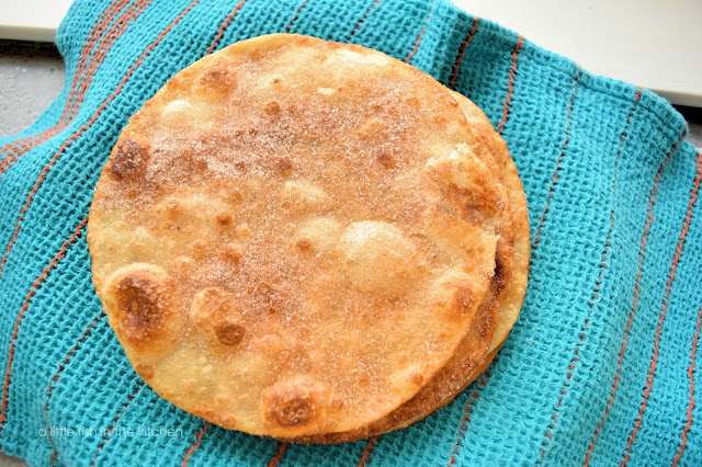 A stack of fresh, crispy buñelos is ready to serve. The crispy fritters are resting on a dark teal tea towel with dark orange stripes.