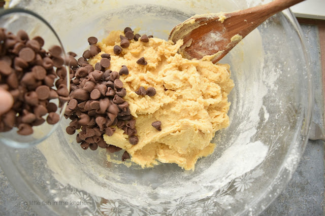 Chocolate chips are mixed into the buttery, rich prepared cookie dough using a wooden spoon.