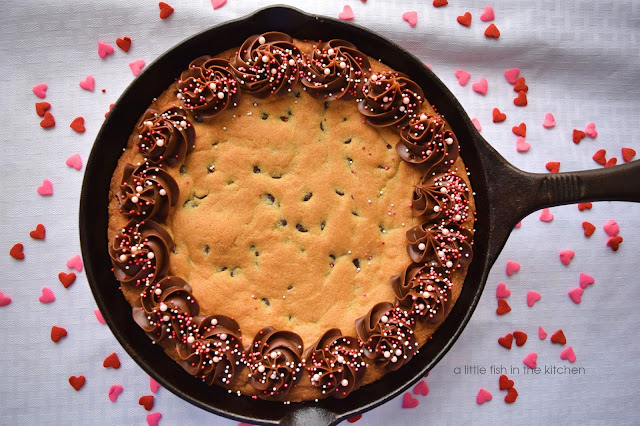 A delicious, fresh baked chocolate chip skillet cookie is topped with swirls of chocolate frosting and pink sprinkles for Valentine's Day.