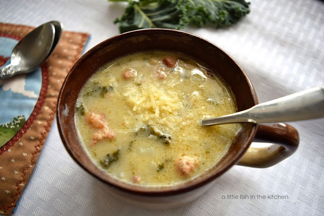 A bowl of creamy Tuscan sausage and white bean soup with kale is served in a rustic brown ceramic soup bowl. The bowl is on a white linen table cloth. Bits of green kale, Italian sausage, white beans and shredded Parmesan cheese are visible on the surface of the warm soup. 