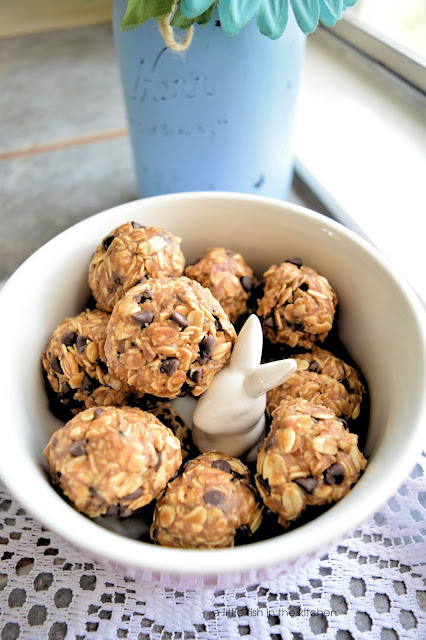 A fresh batch of oat, maple energy bites is ready to enjoy. Hearty oats, sweet chocolate chips and peanut butter are all visible in these no-bake cookies. The energy bites are in a pink ceramic bowl with a white ceraminc bunny in the center. Beside the bowl is a large mason jar that's painted sky blue with pastel  flowers in it.