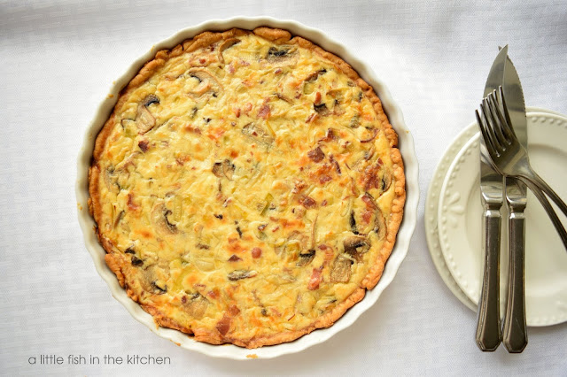 A golden leek and mushroom tart looks inviting in a white ceramic tart dish. Small white appetizer plates are next to the tart with cutlery laying on top. The tart looks tempting with bits of smoky bacon, leek, and mushrooms speckled on it's top. 