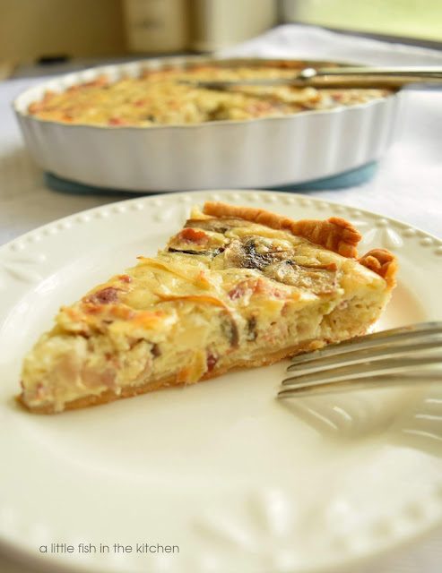 A single slice of this savory leek, bacon and mushroom tart reveals a creamy egg filling dotted with flavorful add-ins. The slice of tart is on a white ceramic plate with a fork resting on the side of the plate. The scalloped white ceramic tart dish with the remainder of the tart is slightly blurred in the background. 