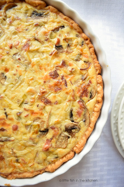 Bits of cheese, tender mushrooms, and bacon are visible in the custardy filling of this flavorful leek, bacon and mushroom tart. The golden pie crust around the rim of the white ceramic tart dish looks crisp and flaky. 