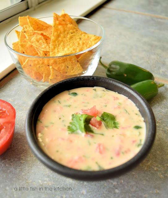 warm white cheese dip is served in a black bowl. Bits of green peppers, and red tomatoes are visible thoughout the cheese dip. The bowl is garnished with a few whole cilantro leaves and chopped tomato. A clear glass bowl filled with tortilla chips, two whole green jalapeno pepper and a sliced whole tomato surround the bowl for decorative effect.
