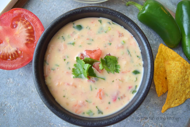 Smooth, warm white cheese dip is served in a black bowl. Bits of green peppers, and red tomatoes are visible thoughout the cheese dip. The bowl is garnished with a few whole cilantro leaves and chopped tomato. Two tortilla chips, two whole green jalapeno pepper and a sliced whole tomato surround the bowl for decorative effect.